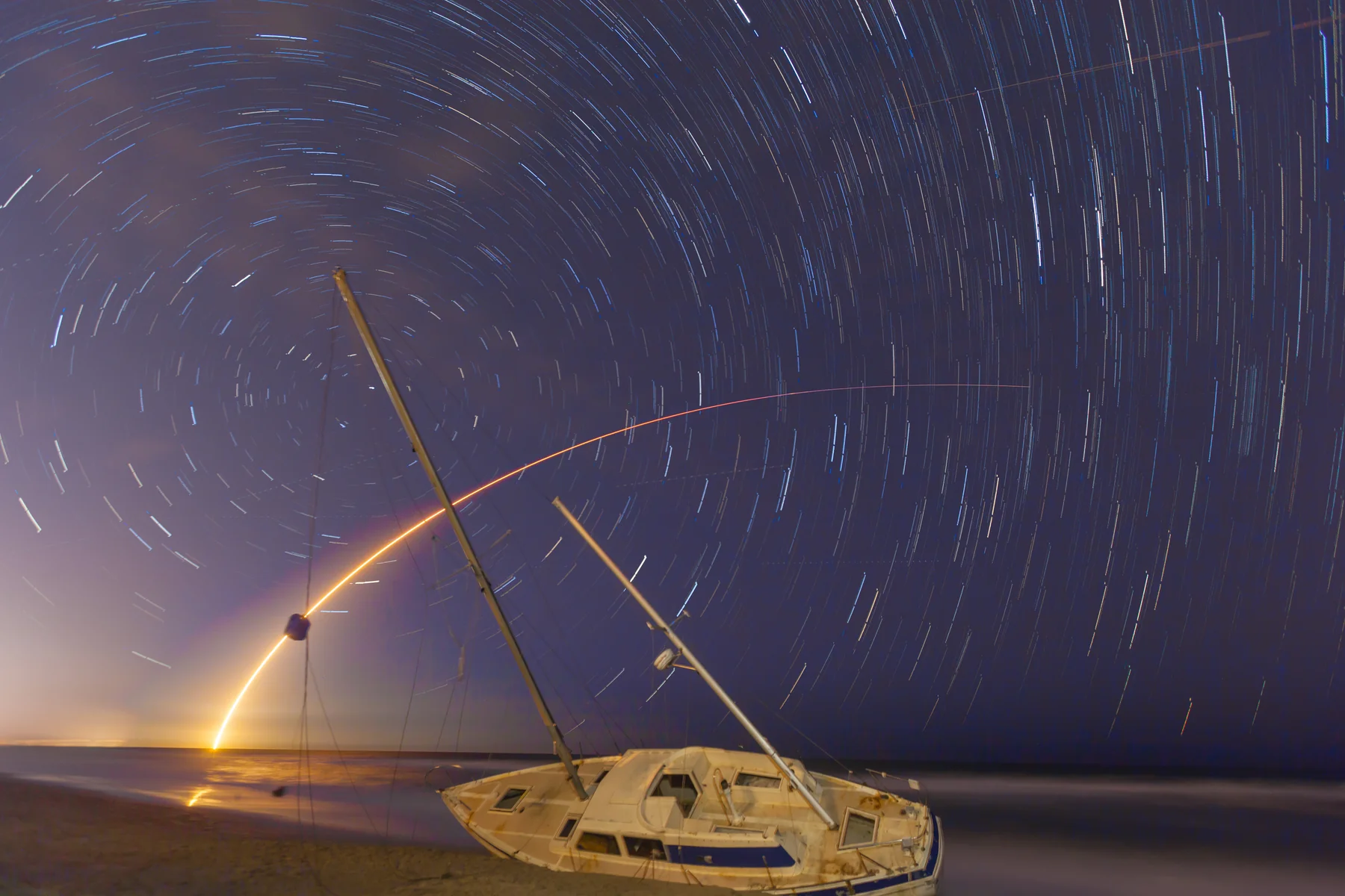 Star trail composite with rocket launch over a beached sailboat on the Space Coast shore