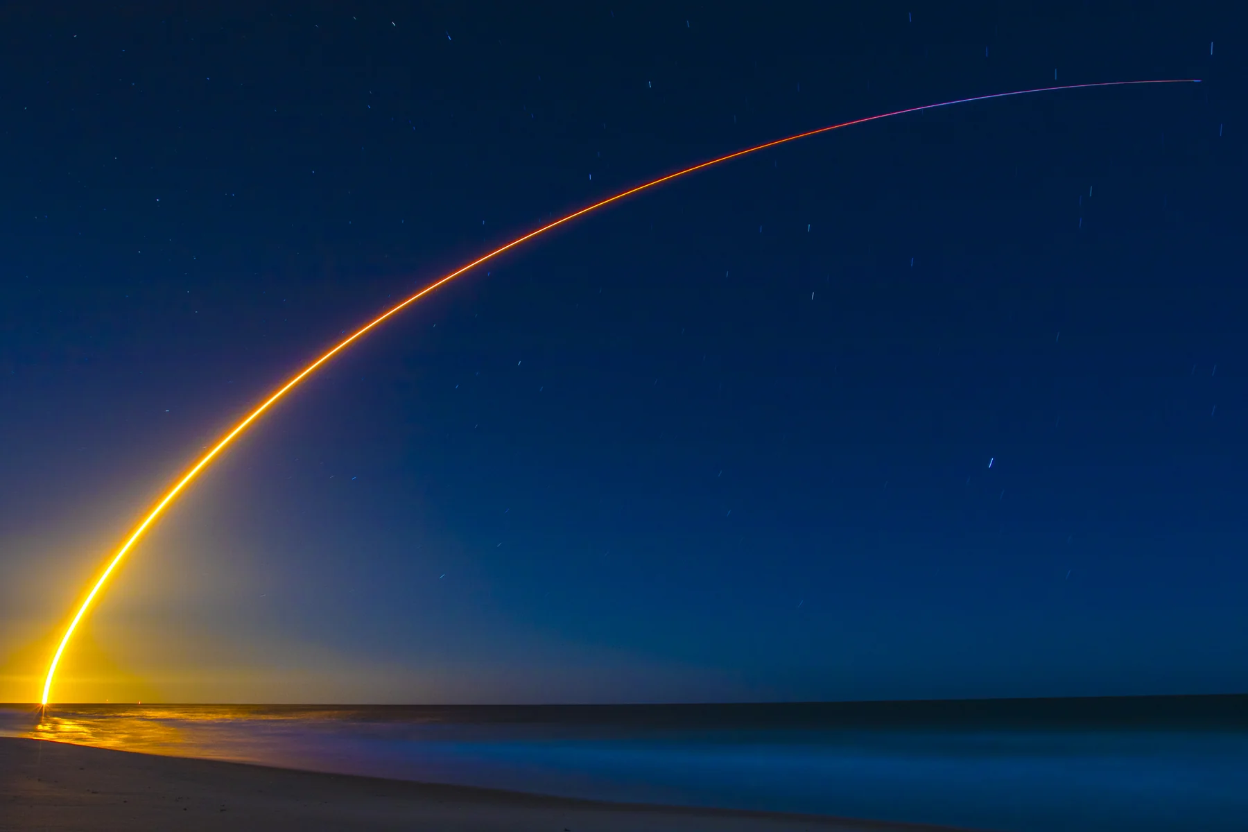 Rocket launch arc over the Space Coast beach at night, golden trail rising into a deep blue sky